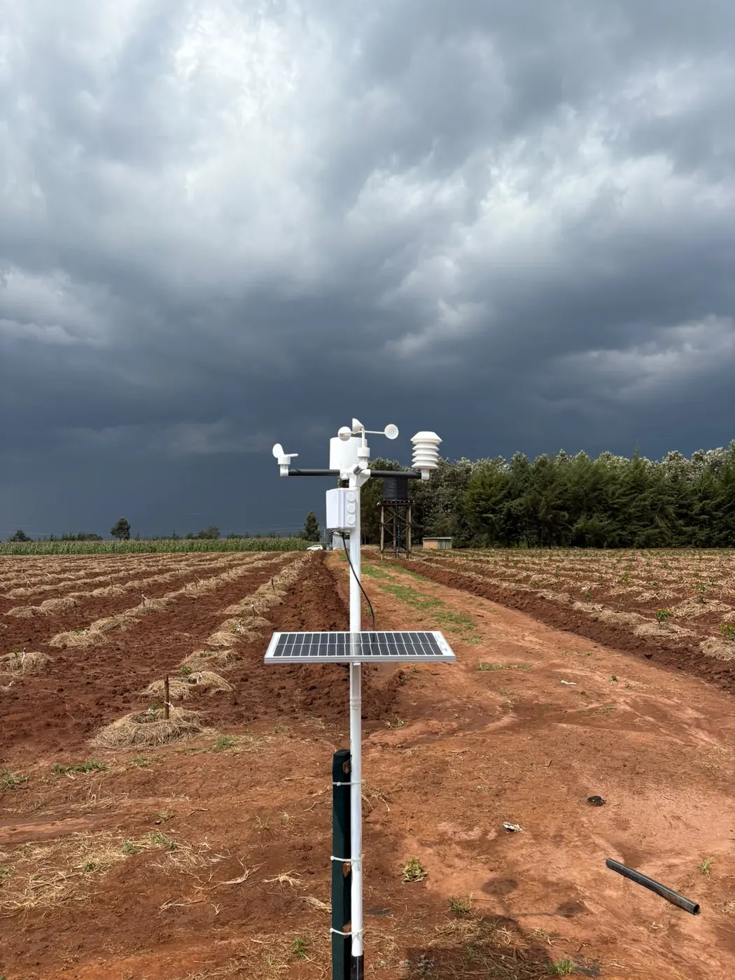 White weather station with wind cups and solar panel on a pole above red ridged farmland under dark storm clouds.